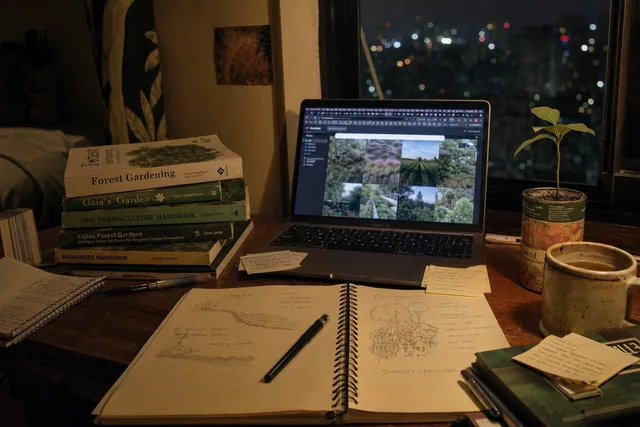 Illustrative image: a desk late at night with a glowing laptop, permaculture books and handwritten garden notes